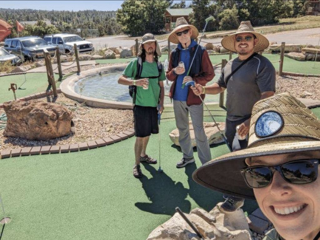 Four friends pose together at a mini-golf course, surrounded by greenery and a small pond.
